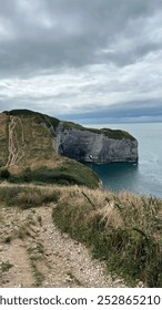 Dramatic coastal cliffs with a rugged path leading along the edge, overlooking the calm ocean. The scenic view is complemented by cloudy skies and natural beauty.