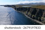 Dramatic coastal cliffs with waterfall flowing into the sea, Isle of Skye, Scotland. Scenic ocean view with rugged rocks, mountains, and blue water under a bright summer sky