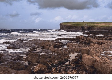 Dramatic coastal landscape with crashing waves, rocky shoreline, and grassy cliff under a moody sky. A wild and untamed view of nature’s raw beauty.