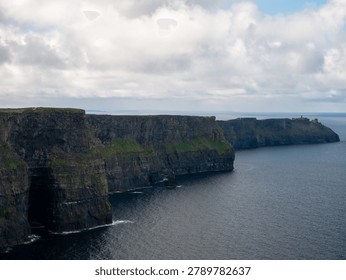A dramatic coastal landscape features towering sea cliffs above a calm ocean under a cloudy sky with distant headlands and rugged rock formations.
