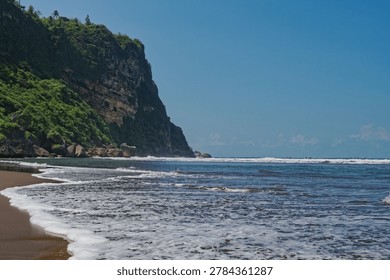 Dramatic coastal landscape featuring a high rocky cliff covered in greenery next to a sandy beach with rolling ocean waves. Ideal for travel, adventure, and real estate backgrounds.