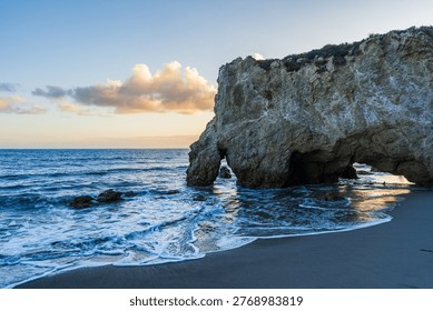 A dramatic coastal landscape featuring a massive limestone rock formation with a natural sea arch and cave carved by ocean waves. Gentle surf washes onto a dark sandy beach at golden hour, with warm sunset clouds reflected in the wet sand. The Pacific Ocean stretches to the horizon under a soft blue and golden sky. This stunning scene captures the rugged beauty of the California coastline, reminiscent of El Matador State Beach in Malibu. Perfect for travel, nature, and outdoor recreation themes.