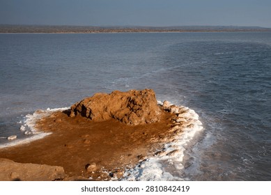 Dramatic coastal landscape featuring a reddish-brown rocky outcrop, white salt deposits, calm blue water, and distant shoreline beneath a clear sky at Rann of Kutch, Gujarat.