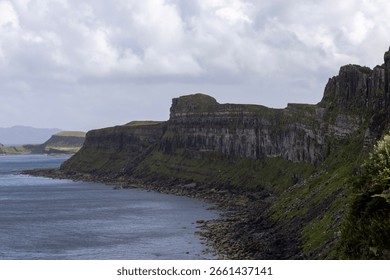 A dramatic coastal landscape featuring steep cliffs covered in lush green vegetation. The rocky shoreline meets calm sea waters under a cloudy sky, creating a wild and untouched natural scene.