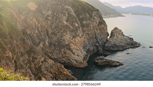 Dramatic coastal landscape featuring steep rocky cliffs and calm ocean waters. Golden hour sunlight creates warm lens flare. Rugged terrain with green vegetation.