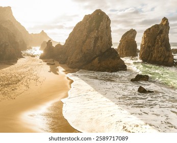 Dramatic coastal landscape featuring towering rock formations, golden sand, and ocean waves crashing against the shore under a warm sunset.  Stock photo