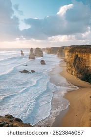 Dramatic coastal landscape featuring towering limestone sea stacks rising from the deep blue ocean alongside a massive sheer cliff face. Rugged rock formations, natural coastal scenery, and powerful ocean waters create a striking geological landscape under a dramatic sky. Ideal for travel, nature, geology, tourism, adventure, and scenic landscape concepts.