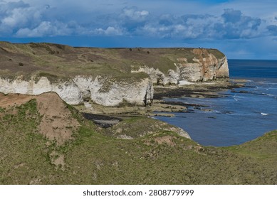 A dramatic coastal landscape featuring white chalk cliffs and a green headland overlooking the blue sea under a cloudy sky at Flamborough, UK.