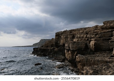 A dramatic coastal scene featuring rugged cliffs overlooking a restless sea under a moody, overcast sky. The textured rock formations contrast with the choppy waters, creating a sense of natural power - Powered by Shutterstock