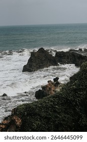 Dramatic coastal scenery featuring powerful ocean waves crashing against dark volcanic rock formations on a rugged shoreline. The foamy white surf contrasts with the textured cliffs and deep green sea