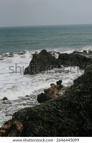 Dramatic coastal scenery featuring powerful ocean waves crashing against dark volcanic rock formations on a rugged shoreline. The foamy white surf contrasts with the textured cliffs and deep green sea