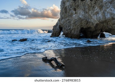 A dramatic coastal seascape featuring a massive limestone sea cave and natural rock arch carved by the relentless Pacific Ocean. Powerful white-capped waves crash and surge across the dark volcanic sand beach, leaving white foam swirling around a weathered piece of driftwood in the foreground. The towering rocky cliff dominates the right side of the frame, its rugged texture contrasting with the smooth wet sand reflecting the warm golden light of the setting sun. Dramatic clouds tinged with pink and gold drift across a pale blue twilight sky on the horizon. The scene evokes a sense of wild, untamed natural beauty, solitude, and the raw power of the ocean. This stunning landscape photograph is ideal for travel, nature, adventure, and environmental themes, suitable for editorial use, wall art, travel brochures, and nature conservation campaigns.