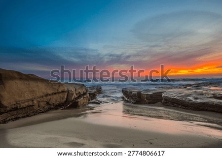 A dramatic coastal sunset in La Jolla, Southern California, where rugged sandstone formations frame a narrow opening to the ocean. The sky glows with vivid hues of orange, pink, and deep blue, reflecting softly across the wet sand. Gentle waves roll through the rocky channel, creating a peaceful yet striking California seascape.