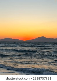 A dramatic coastal sunset scene captures the sun setting behind distant mountain silhouettes over a wide open sea with rolling waves in the foreground. The sky transitions from deep red and vivid orange near the horizon to a soft pale yellow and grey above. Ideal for travel, nature, and inspirational design projects.