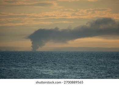A dramatic coastal sunset scene with a thick plume of dark smoke rising in the distance over the deep blue sea, contrasting sharply with the orange and golden tones of the cloudy sky.