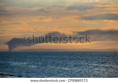 A dramatic coastal sunset scene with a thick plume of dark smoke rising in the distance over the deep blue sea, contrasting sharply with the orange and golden tones of the cloudy sky.