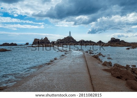 Dramatic coastal view of La Corbiere lighthouse on a rocky island, connected by a tidal causeway under stormy skies. Jersey, Channel Islands, UK.