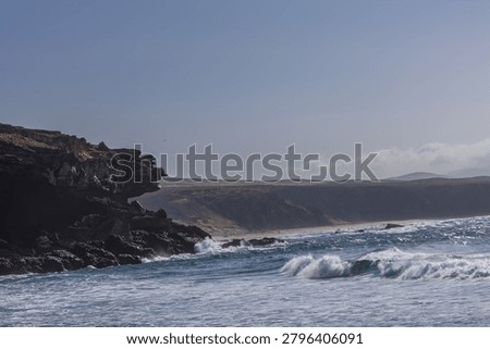 A dramatic coastal vista captures powerful turquoise waves rolling past majestic volcanic cliffs into a hazy desert bay. The atmospheric light and misty mountain horizon create a captivating feeling of untamed freedom, raw nature, and expansive serenity.