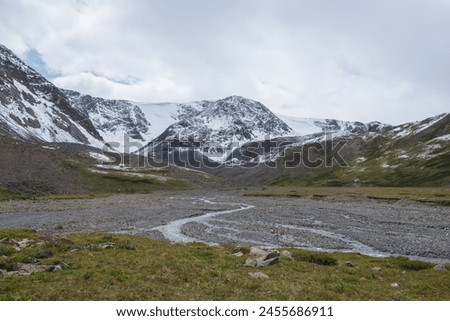 Dramatic cold landscape with river meanders against snow covered large mountain range under rainy cloudy gray sky. Gloomy rainy weather in high mountains. Alpine view to big snowy mount in rain time.