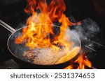 A dramatic cooking scene with a skillet on a gas stove, where large flames are bursting from the pan, indicating high-temperature cooking or flambé technique.