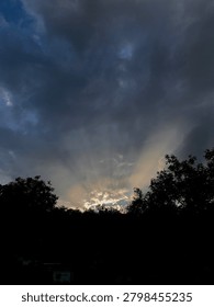 Dramatic crepuscular rays shining through dark storm clouds at sunset. Sunlight streams from behind the cloud cover, illuminating the sky above a silhouette of trees and creating a striking atmospheric landscape.
