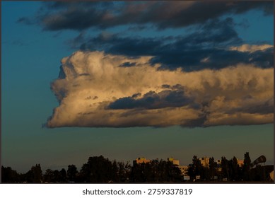 Dramatic cumulus cloud formation illuminated by the setting sun over a city skyline. Large golden cloud hovering above residential buildings and trees at dusk.