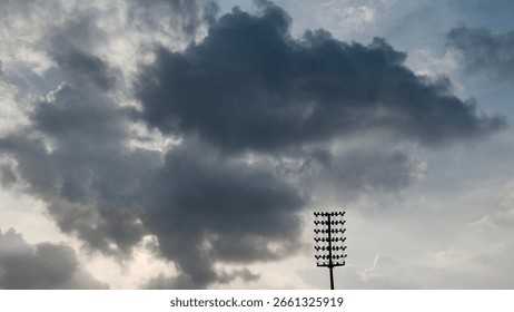 Dramatic dark clouds gather above a tall stadium floodlight, silhouetted against the moody evening sky, signaling the onset of rain or a sports event