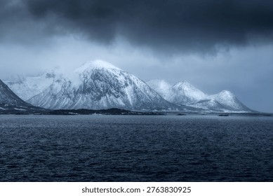 Dramatic dark clouds gather above majestic snowy peaks in Fiskebol, Lofoten, Norway. The tranquil waters reflect the icy mountains, creating a breathtaking view. The cold air carries the promise of winter adventures, inviting exploration of this serene Arctic paradise.