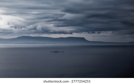 Dramatic dark clouds loom over calm, deep blue water with distant layered mountains and a tiny island.