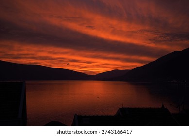 Dramatic dark orange sunset over a quiet bay with mountain silhouettes and fiery twilight clouds reflecting on the water.