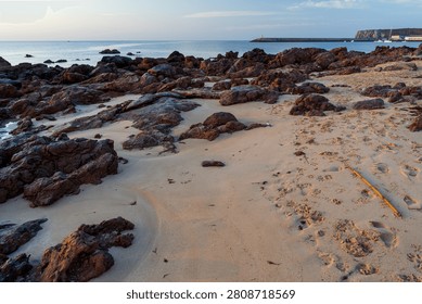 Dramatic dark red rocks emerge from golden beach sand at low tide along the coastline near Sagres in Portugal. A harbour breakwater with a lighthouse and a dramatic headland rises softly in the distant evening haze.