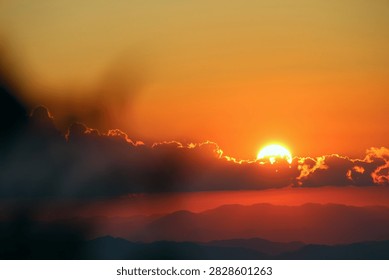 Dramatic dark red sunset with the bright sun hiding behind dark clouds over mountain silhouettes. Stunning deep red and black sunset sky with glowing sun behind dark clouds. 