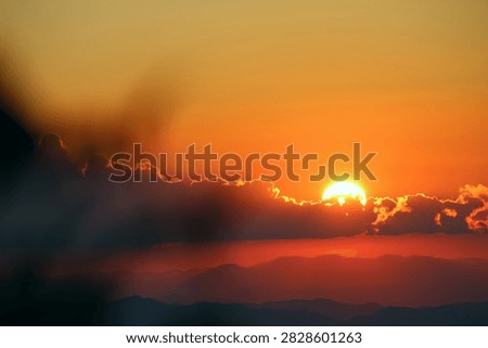 Dramatic dark red sunset with the bright sun hiding behind dark clouds over mountain silhouettes. Stunning deep red and black sunset sky with glowing sun behind dark clouds. 