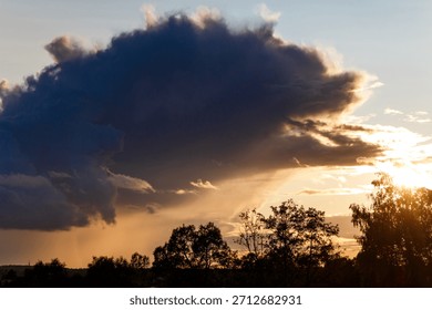 Dramatic dark storm cloud looms over silhouetted trees during a fiery summer sunset with rays of light bursting through the horizon