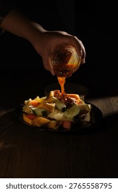 A dramatic dark studio shot of a hand pouring thick, glossy Indonesian rujak sauce from a glass bowl over a generous mound of assorted fresh tropical fruit pieces on a dark plate, with the amber-orange sauce cascading in a continuous stream that catches the dramatic single light source against a near-black background. The colorful fruit pieces including cucumber, papaya, mango, jicama, and pineapple are illuminated in warm tones, creating a visually striking and cinematic food photography composition.