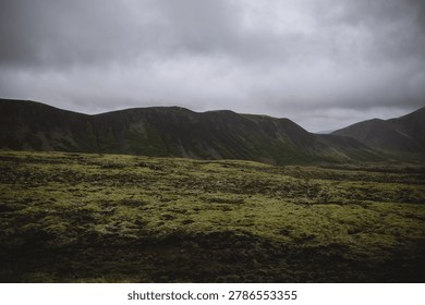 Dramatic dark volcanic mountain ridge rising above a wide, vibrant green moss field under a heavy, brooding overcast sky, showing untamed wilderness and raw natural power.