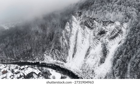 Dramatic drone scene of a mist wrapped escarpment towering above a river and small settlement, snowy folds etched into the slope. Atmospheric winter landscape merging geology, wilderness, and quiet village life.