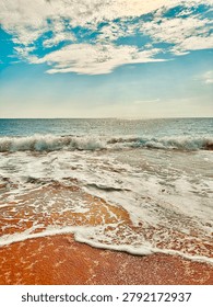 A dramatic and energetic beach scene featuring powerful waves crashing onto a golden-brown sandy shoreline, producing rich white foam that spreads dynamically across the wet sand. The sky above is filled with dramatic alto and cirrus cloud formations against a vivid blue backdrop, creating a sense of movement and atmospheric energy. The composition captures the raw power and beauty of tropical coastal waves with strong contrast between the warm reddish-golden sand, pure white foam, and brilliant blue sea and sky