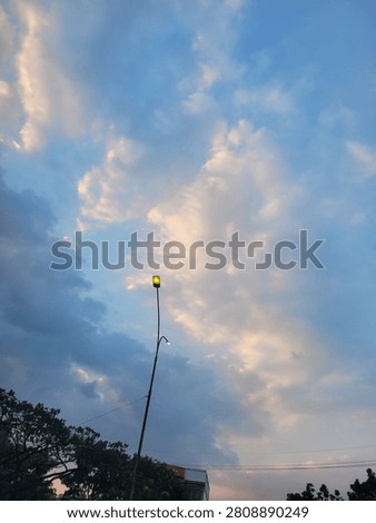 Dramatic evening clouds illuminated by soft sunset light above an urban street in Bandung, West Java, Indonesia. A glowing street lamp contrasts beautifully with the blue sky, creating a peaceful cityscape atmosphere before nightfall.