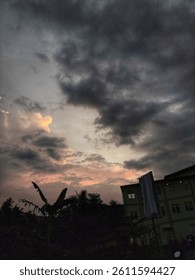 Dramatic evening sky with dark clouds and glowing sunset light over silhouettes of trees and buildings in a tropical urban area.

