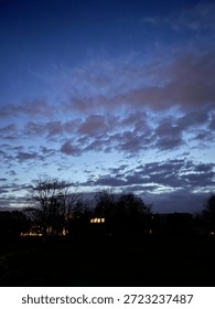 Dramatic evening sky with dark clouds at dusk over silhouetted residential buildings and bare trees. Deep blue twilight atmosphere with illuminated windows glowing in the darkness. Moody cloudscape during blue hour with contrasting light and shadow.