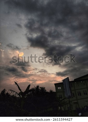 Dramatic evening sky with dark clouds and glowing sunset light over silhouettes of trees and buildings in a tropical urban area.
