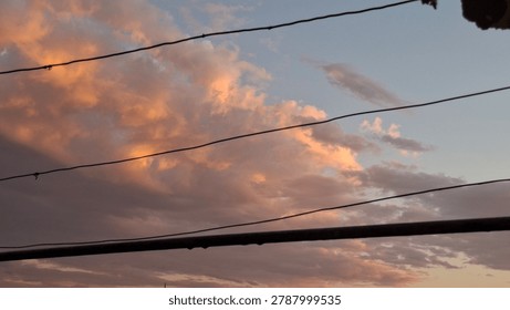 Dramatic evening sky filled with glowing pink and orange clouds during sunset, captured from an urban residential area with visible utility wires crossing the composition. The warm pastel light illuminates textured cloud formations against a fading blue-gray sky, creating a cinematic and atmospheric twilight mood. Dark silhouetted cables and balcony elements frame the colorful sunset scene, adding contrast and a realistic city perspective to the natural sky display. The image emphasizes changing weather, peaceful dusk atmosphere, and the beauty of soft evening light after sunset. Rich cloud textures and warm tones create a visually emotional landscape suitable for backgrounds, weather concepts, urban lifestyle visuals, dramatic sky themes, and inspirational nature content. The balance between urban infrastructure and vibrant natural colors makes the composition ideal for editorial, atmospheric, environmental, and seasonal evening imagery.