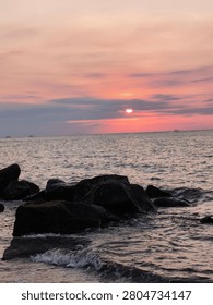 A dramatic evening sky with soft clouds partially obscuring the setting sun over the open sea. The foreground features water splashing gently against wet, dark boulders on the beach.