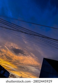 Dramatic evening sky at sunset with gradient colors of deep blue, purple, orange and yellow. Silhouette of overhead electric wires, street lamps and building outlines in a residential urban area. Natural light, atmospheric view, no visible legible text or brand logos.