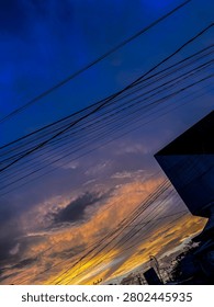 Dramatic evening sky at sunset with gradient colors of deep blue, purple, orange and yellow. Silhouette of overhead electric wires, street lamps and building outlines in a residential urban area. Natural light, atmospheric view, no visible legible text or brand logos.