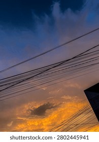 Dramatic evening sky at sunset with gradient colors of deep blue, purple, orange and yellow. Silhouette of overhead electric wires, street lamps and building outlines in a residential urban area. Natural light, atmospheric view, no visible legible text or brand logos.