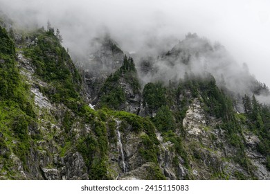 Dramatic fog over jagged rocky cliffs with dark green trees, foliage, and a small waterfall - Lake 22, Baker-Snoqualmie National Forest, Washington - Powered by Shutterstock