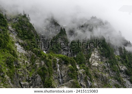 Dramatic fog over jagged rocky cliffs with dark green trees, foliage, and a small waterfall - Lake 22, Baker-Snoqualmie National Forest, Washington