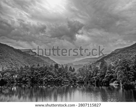 A dramatic forested valley bisected by a calm lake reflects towering clouds and layered mountains under moody overcast skies in the Scottish Highlands.